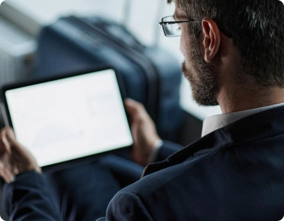 A man in a suit, seen from behind, holds and looks at a tablet. A suitcase is visible in the background, suggesting he is in a travel or business setting—perhaps reviewing reports from an enterprise QA services provider or coordinating with a QA outsourcing company on important project updates. Tristha Global — BFSI QA Services Company & Test Automation